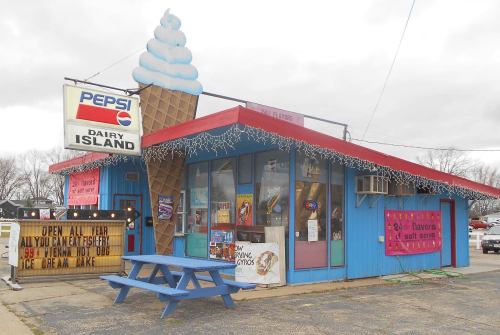A colorful ice cream shop with a large sign, picnic table, and festive decorations, advertising various flavors and treats.