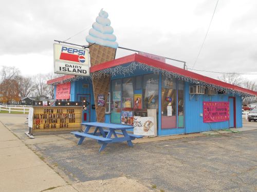 A colorful ice cream shop with a large ice cream cone sign, featuring a picnic table outside and various signs.