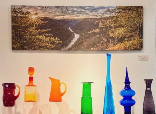 A colorful display of glass vases and pitchers in front of a scenic landscape photograph featuring a river and bridge.
