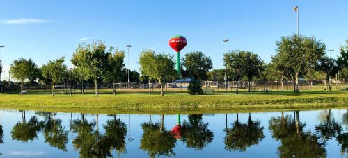 A serene park scene with a water tower and trees reflected in a calm pond under a clear blue sky.
