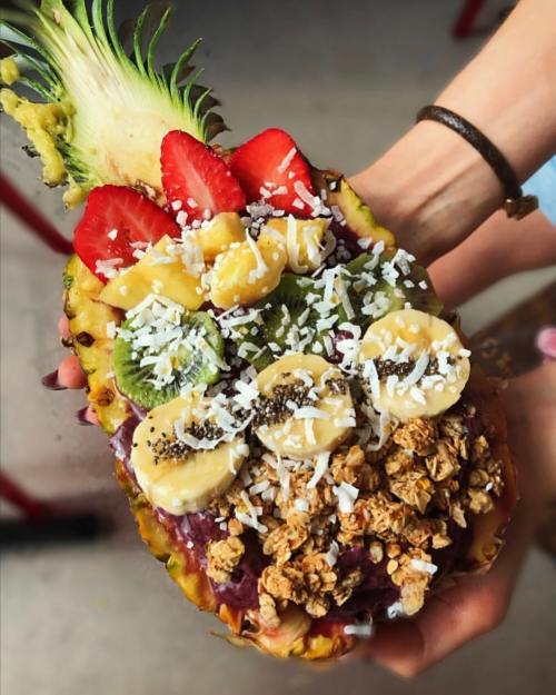 A halved pineapple filled with smoothie, topped with strawberries, bananas, kiwi, coconut, and granola.