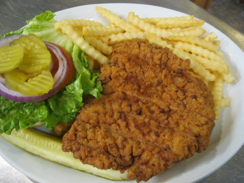 A plate of crispy fried chicken with crinkle-cut fries, lettuce, tomato, onion, pickles, and a cucumber slice.