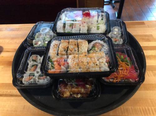A variety of sushi rolls and sashimi arranged in black takeout containers on a wooden table.