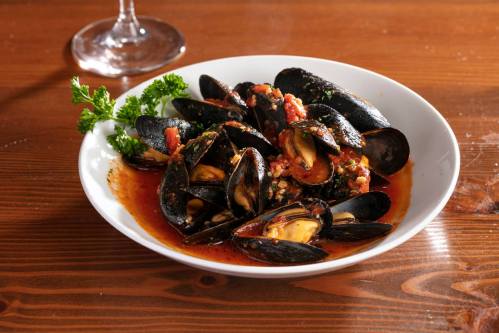 A white bowl filled with mussels in a rich tomato sauce, garnished with parsley, on a wooden table.
