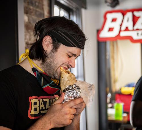 A person wearing a bandana bites into a large burrito, with a restaurant sign in the background.
