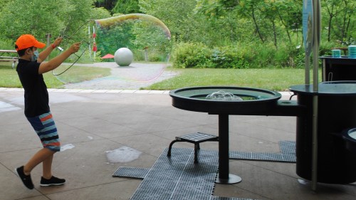 A child in an orange hat creates a large bubble at an outdoor play area with greenery in the background.