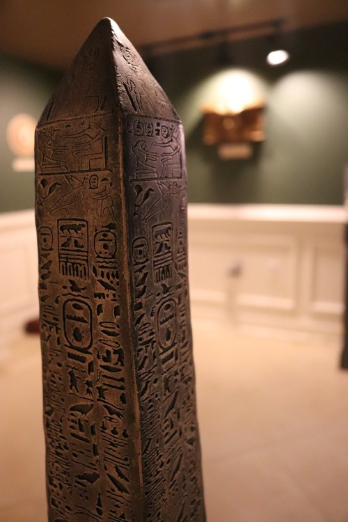 A close-up of an ancient obelisk covered in hieroglyphics, set in a museum with a blurred background.