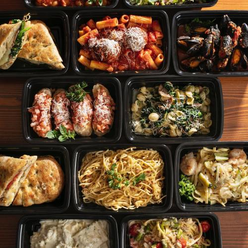 A variety of pasta dishes and seafood in black takeout containers, garnished with herbs on a wooden table.