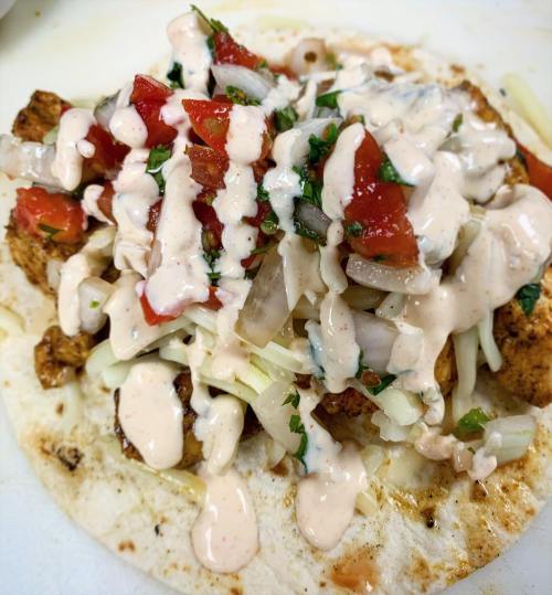 A close-up of a tortilla topped with grilled chicken, diced tomatoes, onions, cilantro, and drizzled with sauce.