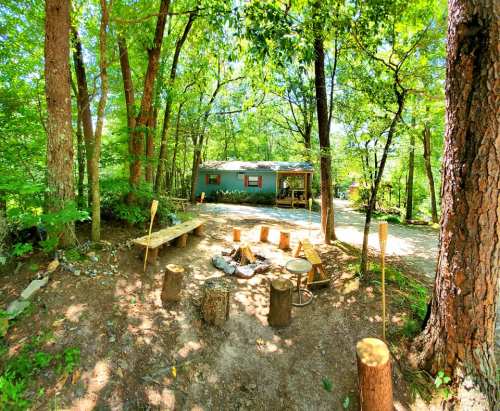 A cozy outdoor scene with a fire pit, wooden seating, and a cabin surrounded by lush green trees.