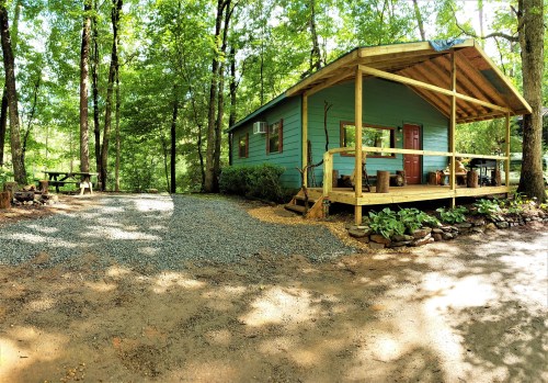 A cozy blue cabin surrounded by trees, featuring a porch and gravel driveway, set in a serene wooded area.