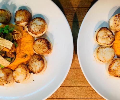 Two plates of seared scallops arranged around a bed of orange puree, garnished with greens and vegetables.