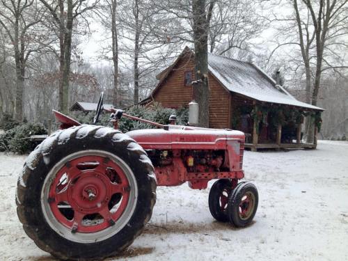 A red tractor covered in snow, with a rustic cabin and trees in the background, creating a winter scene.