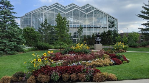 A greenhouse surrounded by vibrant flowers and greenery, featuring a statue in a colorful garden.