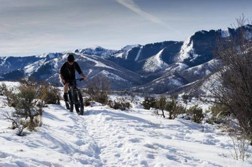 A person rides a fat bike through snowy terrain with mountains in the background under a clear sky.