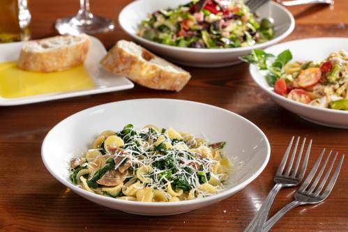 A bowl of pasta with greens and cheese, accompanied by a salad, bread, and a plate of pasta on a wooden table.