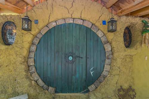 A round wooden door framed by rustic stone, set against a textured yellow wall with lanterns on either side.
