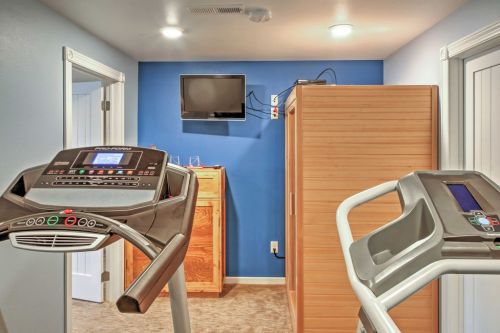 A home gym with two treadmills, a wooden storage unit, and a wall-mounted TV against a blue wall.