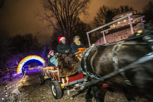 A horse-drawn wagon with passengers rides under a colorful arch of lights in a snowy landscape at night.