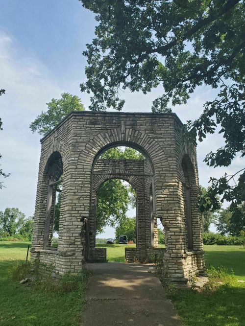 Stone structure with arches surrounded by greenery under a clear sky. Pathway leads to the entrance.
