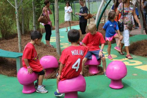 Children in red shirts play on pink mushroom-shaped seats in a park, while adults walk nearby.