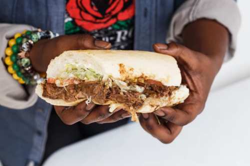 A person holding a sandwich filled with shredded meat, lettuce, and tomato, showcasing vibrant bracelets on their wrist.