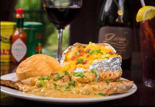 A plate of grilled chicken with shrimp in sauce, a baked potato topped with cheese, a biscuit, and a glass of red wine.