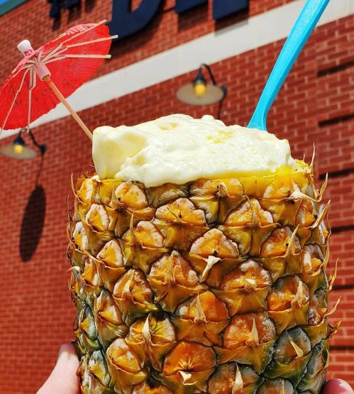 A hand holds a pineapple filled with creamy dessert, topped with a small umbrella and a blue spoon.