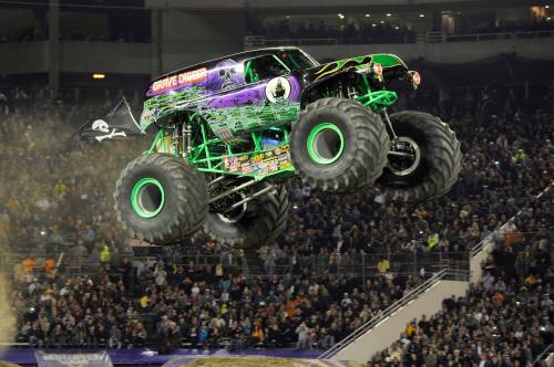 A monster truck with a green and purple design jumps over a crowd at a stadium event, kicking up dust.