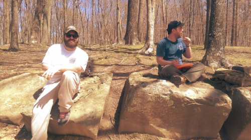 Two men sitting on large rocks in a wooded area, enjoying snacks and the outdoors on a sunny day.