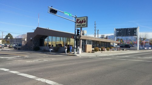 A coffee shop at an intersection, featuring a large sign and a modern building with windows, under a clear blue sky.