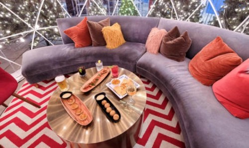 Cozy seating area with colorful pillows, a round table featuring snacks and drinks, and festive lights in the background.