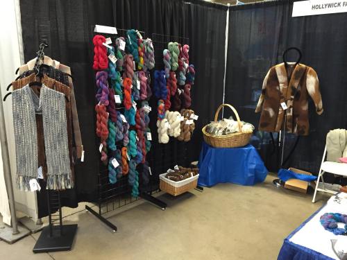 A display of colorful yarn skeins, knitted garments, and a basket of fiber at a craft fair booth.