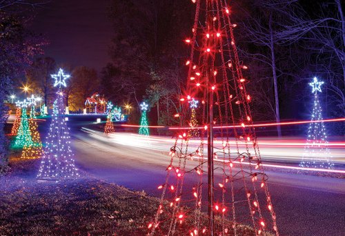 Colorful Christmas trees adorned with lights line a road at night, with car lights creating streaks in the background.