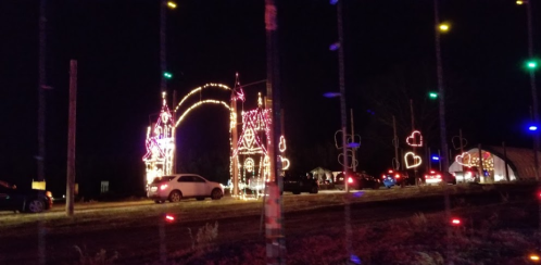 Colorful holiday lights illuminate an archway and heart shapes at a nighttime festive display with cars nearby.