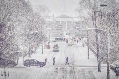 A snowy street scene with cars and pedestrians navigating through heavy snowfall and reduced visibility.