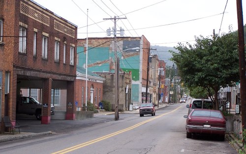 A quiet street scene featuring brick buildings, parked cars, and power lines under a cloudy sky.
