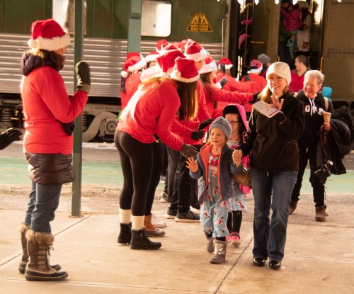 A joyful scene of children and adults in Santa hats, celebrating and greeting each other at a festive event.