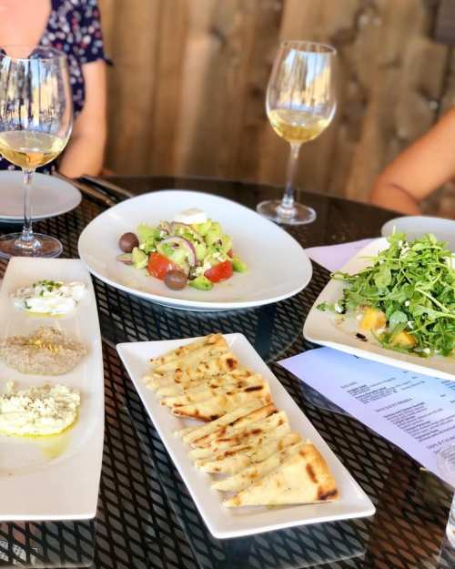 A table set with glasses of white wine and three plates of fresh salads and flatbreads in a sunny outdoor setting.