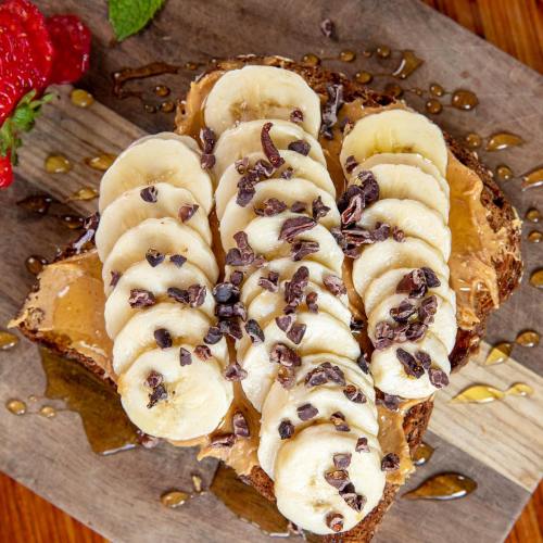 A slice of whole grain toast topped with peanut butter, banana slices, cacao nibs, and a drizzle of honey.