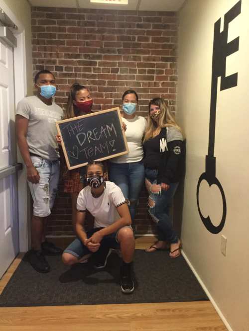 A group of five people poses together in a hallway, holding a sign that says "THE DREAM TEAM!" while wearing masks.
