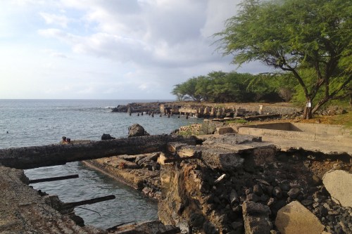 A rocky shoreline with remnants of a pier, surrounded by trees and calm water under a cloudy sky.