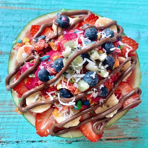 A colorful fruit bowl with strawberries, blueberries, bananas, and toppings, drizzled with chocolate on a green background.