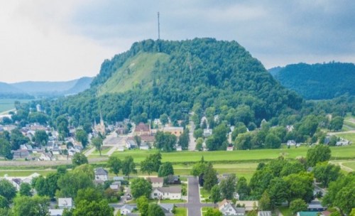 Aerial view of a small town with a prominent green hill and communication tower in the background.