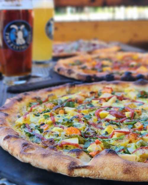 A close-up of a freshly baked pizza topped with vegetables, alongside two glasses of beer in the background.