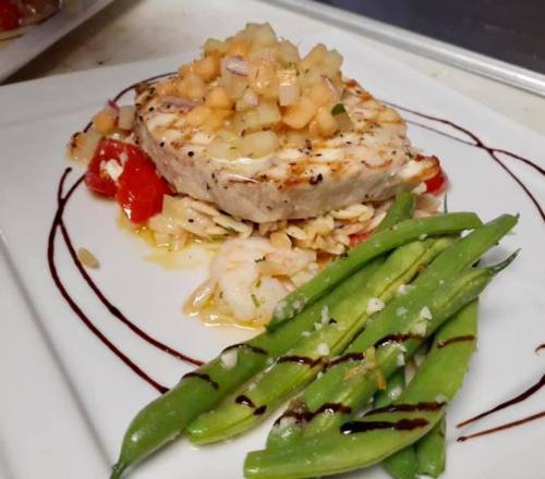 Grilled fish topped with fruit salsa, served with shrimp, tomatoes, and green beans on a white plate.