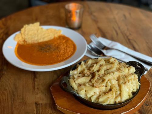 A skillet of creamy macaroni and cheese beside a plate of tomato soup with a crispy bread topping on a wooden table.