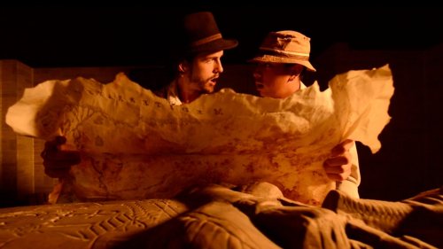 Two men in hats closely examine an ancient map, illuminated by warm light, with a carved stone figure in the foreground.