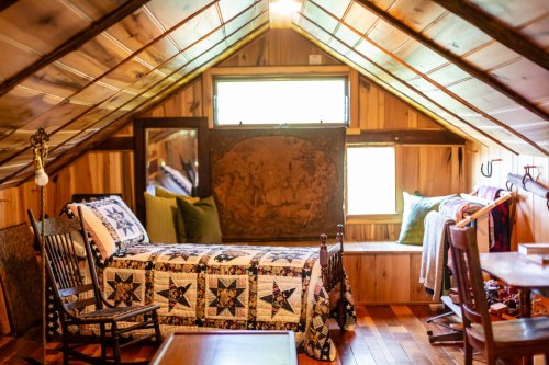 Cozy attic room with a quilted bed, wooden chairs, and natural light from a window, featuring rustic decor.