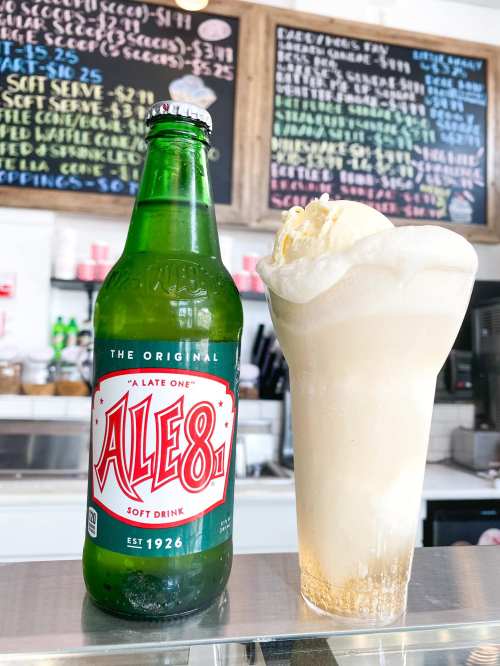 A green bottle of Ale-8 soda next to a frosty glass of ice cream float, with a chalkboard menu in the background.